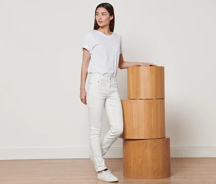 Une femme se tient debout et s'appuie contre une pile de trois socles en bois. Elle porte un t-shirt blanc et un jean Colored Denim « Fit Emma » blanc crème.