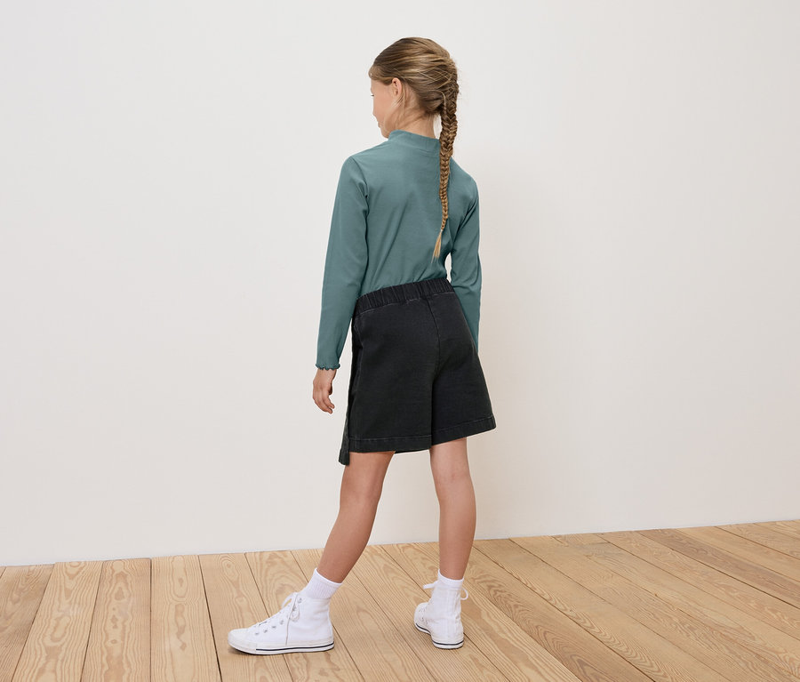 Une fille, vue de dos, porte un t-shirt vert à manches longues, un short noir et des baskets blanches.