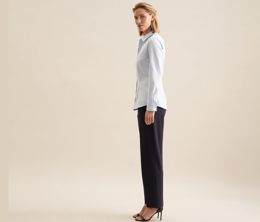 Femme de profil, regardant par-dessus son épaule, portant un chemisier en tulle Seidensticker bleu clair, un pantalon foncé et des sandales noires à talons hauts.
