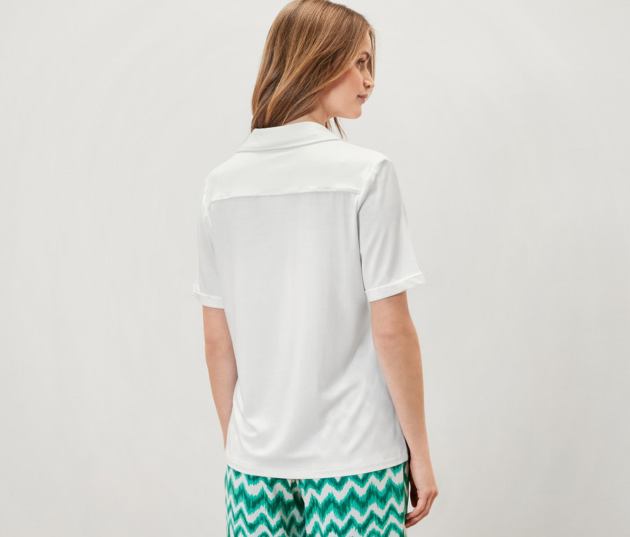 Femme debout, dos à la caméra, portant une blouse blanche avec patte de boutonnage et un pantalon vert à motifs.