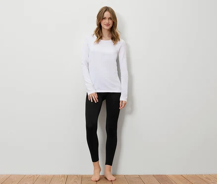 Une femme se tient devant un mur blanc, portant un t-shirt blanc à manches longues et un legging noir.