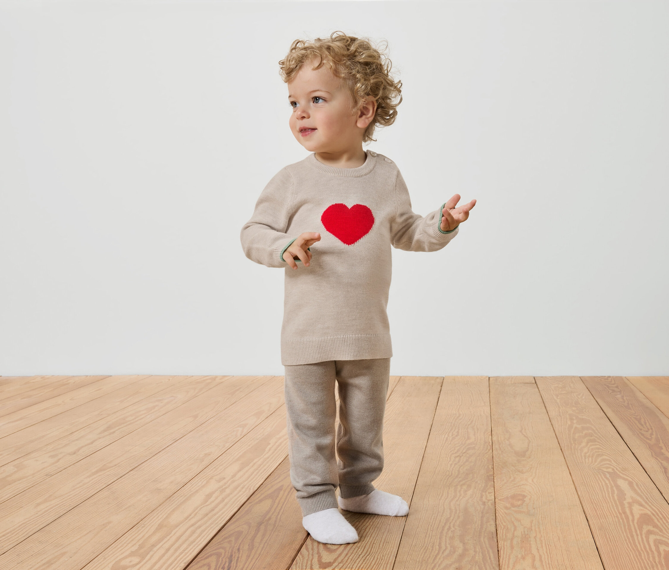 Un bambin aux cheveux bouclés se tient sur un plancher en bois, portant un pull beige avec un cœur rouge et un pantalon beige.