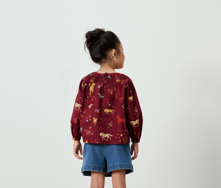 Vue arrière d'une fillette avec un chignon, portant un chemisier bordeaux imprimé de chevaux et un short en jean bleu.