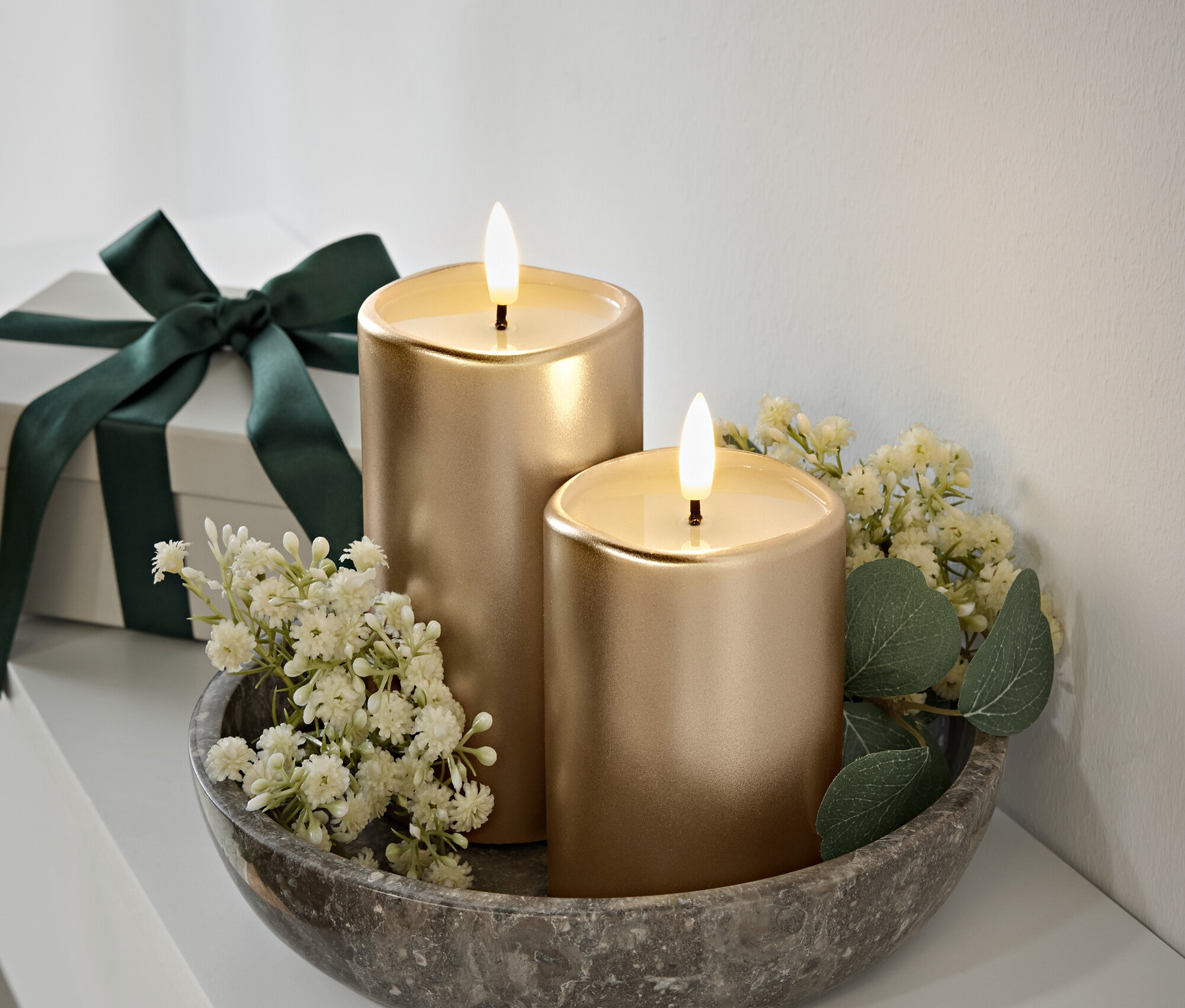Deux bougies dorées avec des flammes dans un bol en pierre grise, des fleurs blanches et des feuilles vertes. Boîte cadeau avec un ruban vert foncé en arrière-plan.