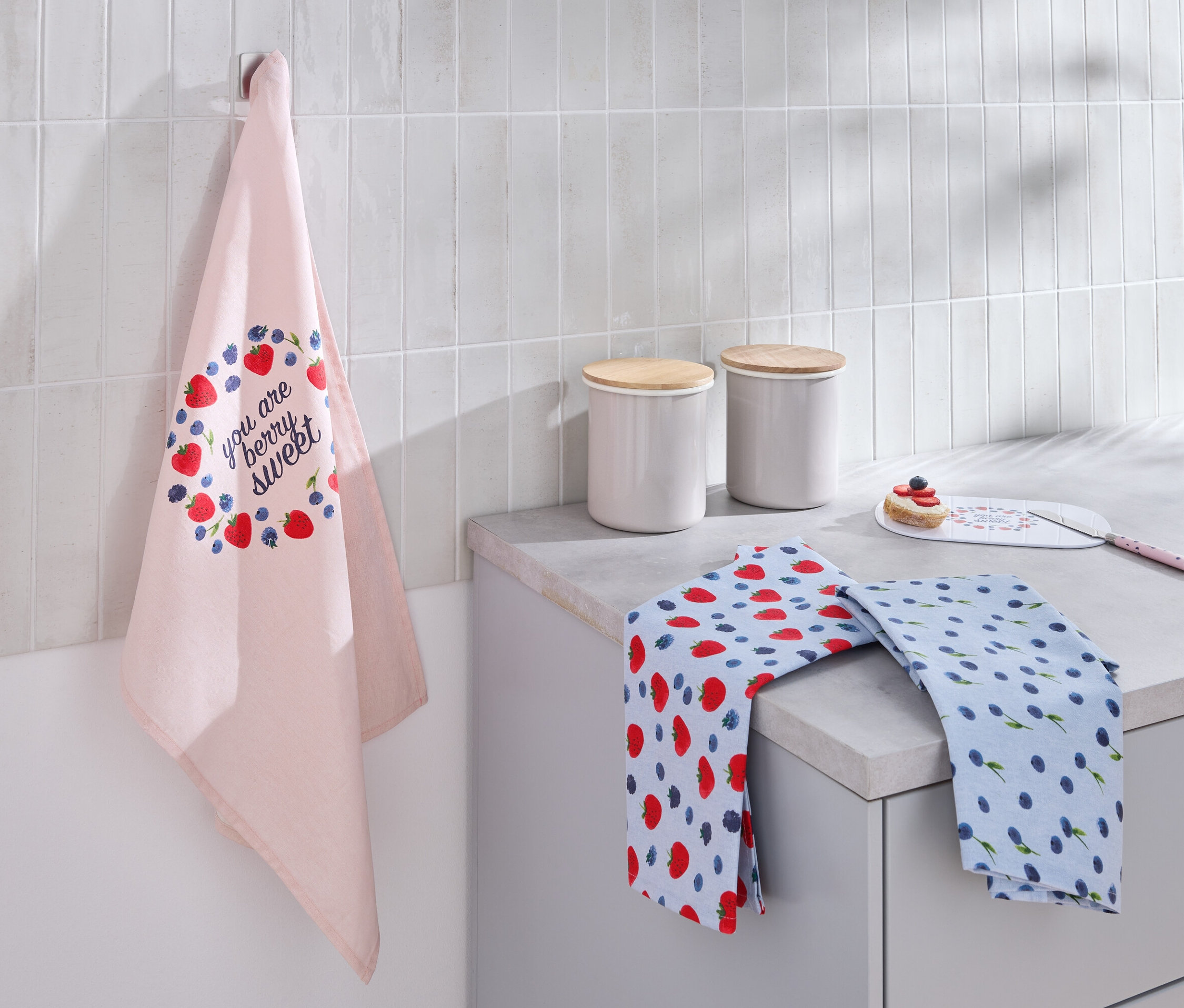 Un torchon rose clair avec un imprimé de fraises et de myrtilles est accroché au mur. Deux torchons pliés et deux bidons blancs avec des couvercles en bois sont posés sur le plan de travail.