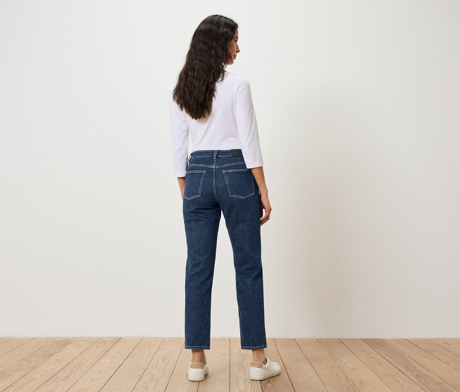 Une femme aux cheveux foncés se tient dos à la caméra, portant un jean bleu et un t-shirt blanc.
