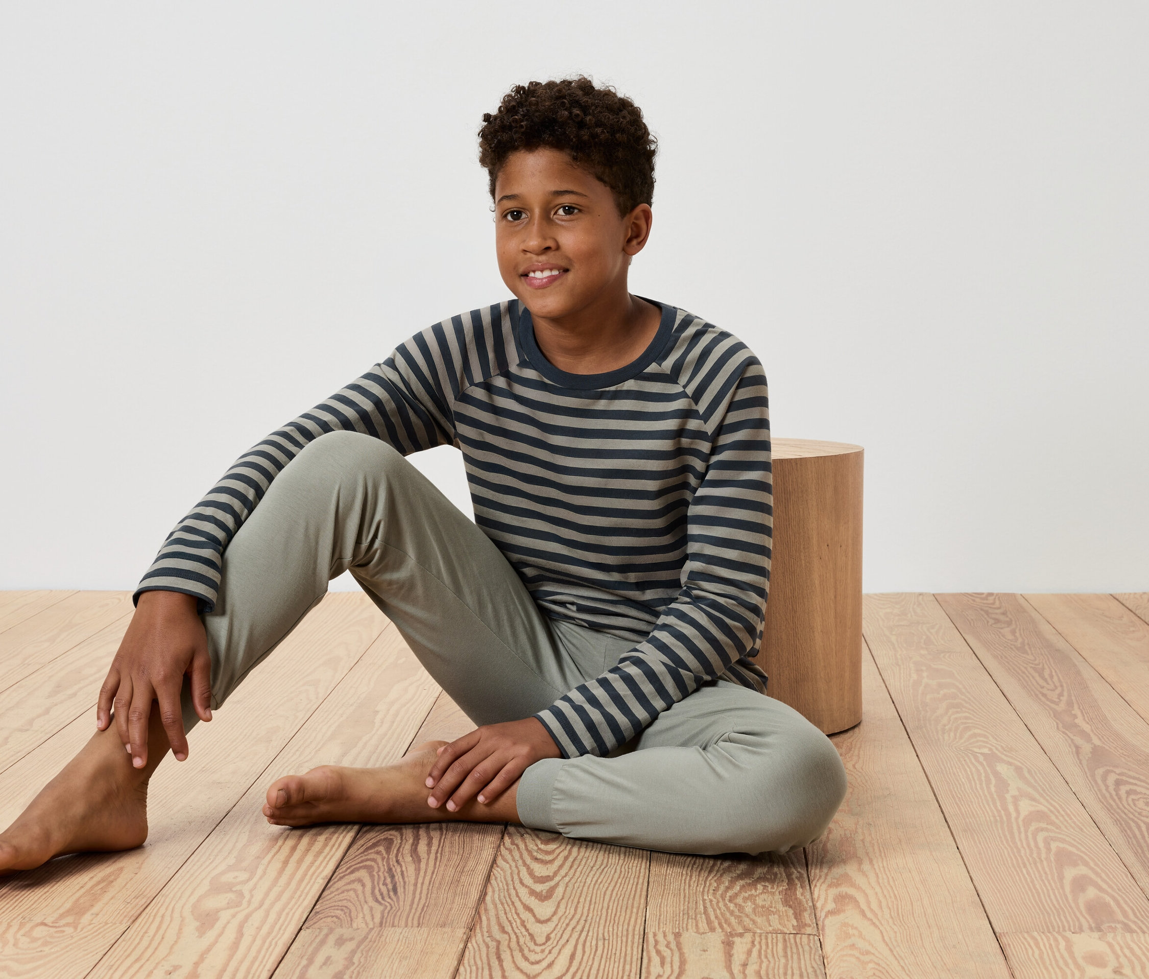 Un jeune garçon est assis sur un plancher en bois, appuyé contre une petite table en bois, portant un t-shirt rayé et un pantalon.
