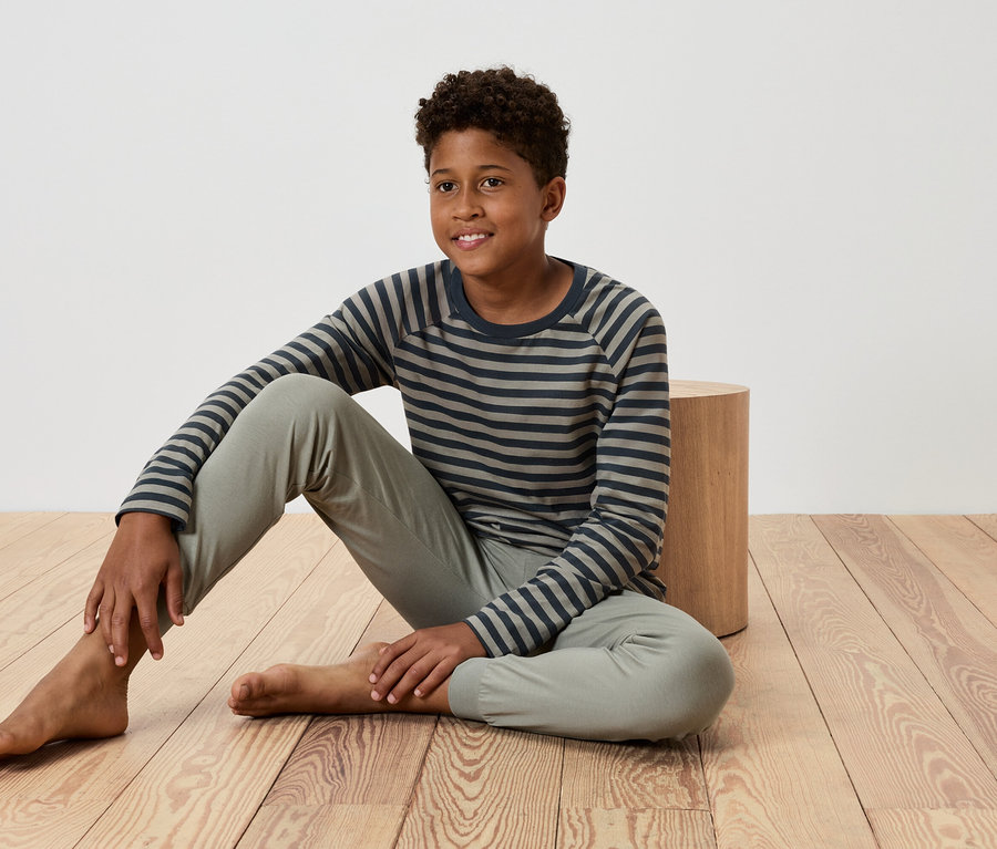 Un jeune garçon est assis sur un plancher en bois, appuyé contre une petite table en bois, portant un t-shirt rayé et un pantalon.