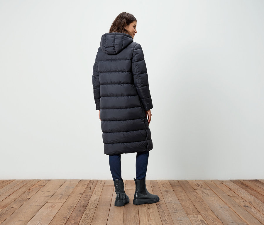 Une femme vue de dos porte un long manteau noir en duvet, un jean et des bottes.