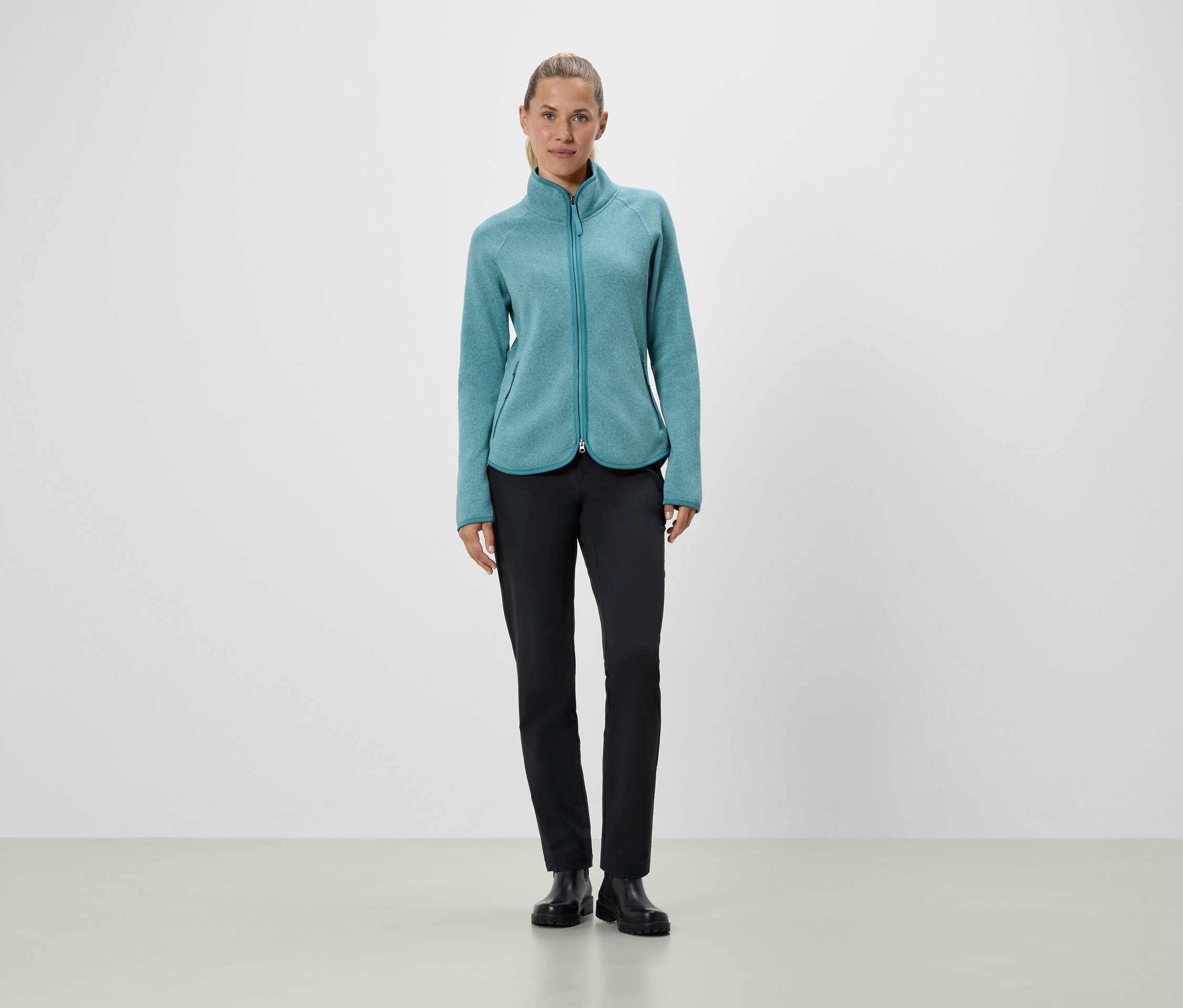 Une femme debout portant une veste en polaire turquoise et un pantalon noir.