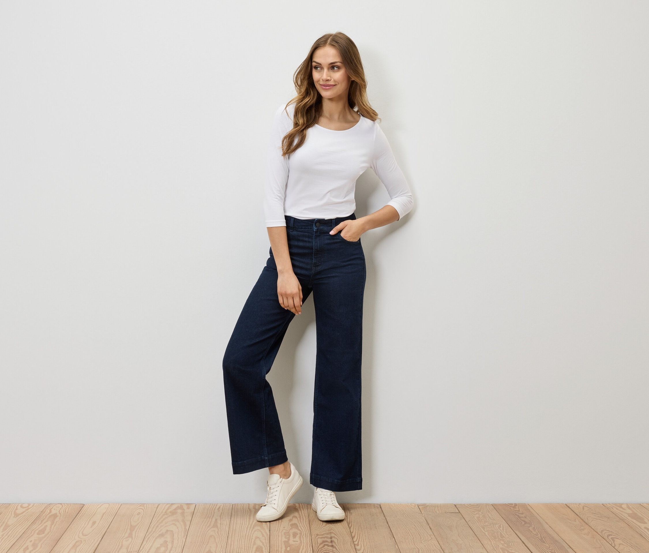 Une femme pose dans un t-shirt blanc à manches trois-quarts, un jean évasé bleu foncé et des baskets blanches.