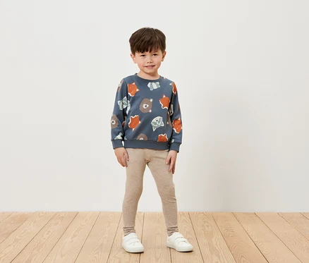 Un garçon portant un sweat-shirt bleu imprimé avec des motifs d'ours et de renards, des leggings beiges et des baskets blanches se tient sur un plancher en bois devant un mur blanc.