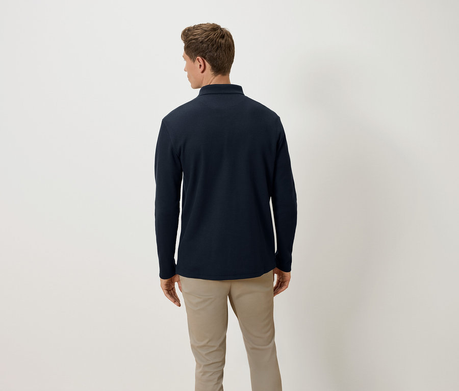 Un homme, dos à la caméra, porte un polo à manches longues bleu marine et un pantalon beige.