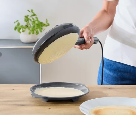Une personne incline une poêle à frire électrique gris foncé au-dessus d'une assiette contenant le reste de la pâte. Une crêpe finie repose sur une assiette blanche sur un comptoir en bois.