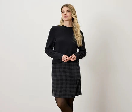 Une femme blonde porte un pull noir et une jupe gris et noir.