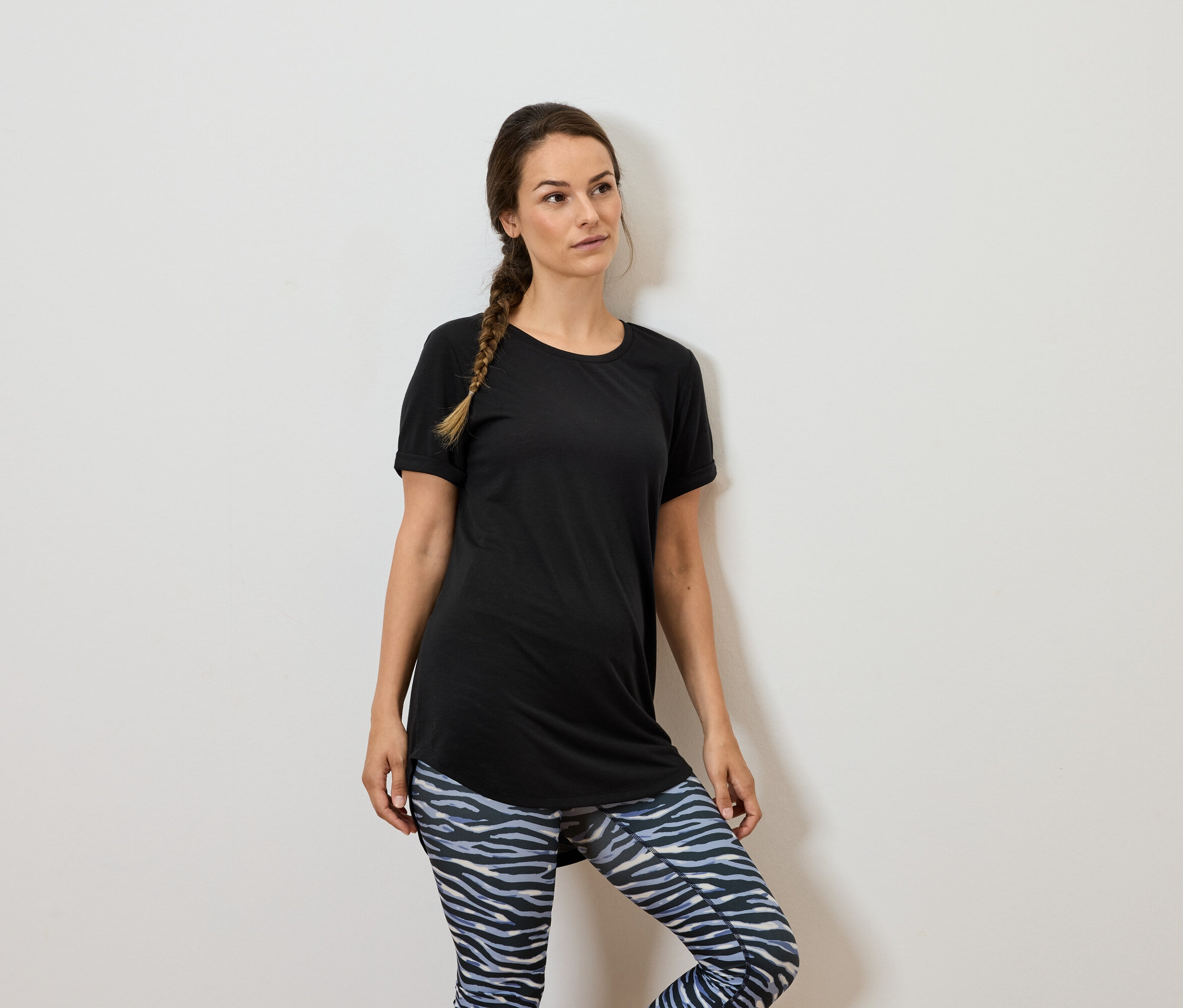 Femme aux cheveux bruns portant un t-shirt noir et des leggings à motifs, debout contre un mur blanc.