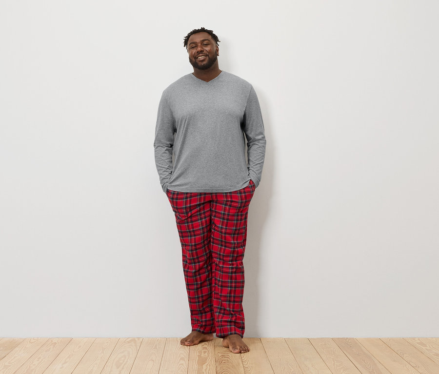 Un Afro-Américain se tient debout, les mains dans les poches, portant un t-shirt gris à manches longues et un pantalon de pyjama à carreaux rouges.
