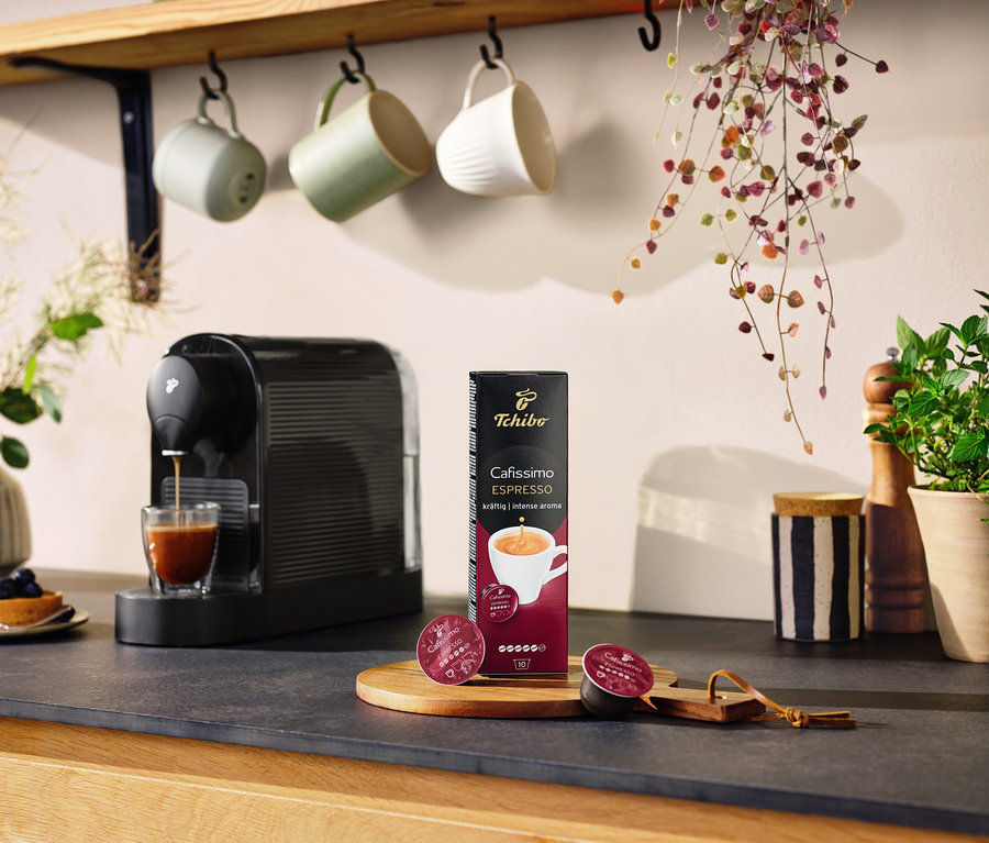 Une Cafissimo pure+ noire prépare du café dans un verre. Un paquet de capsules et deux capsules sont posés sur la table.