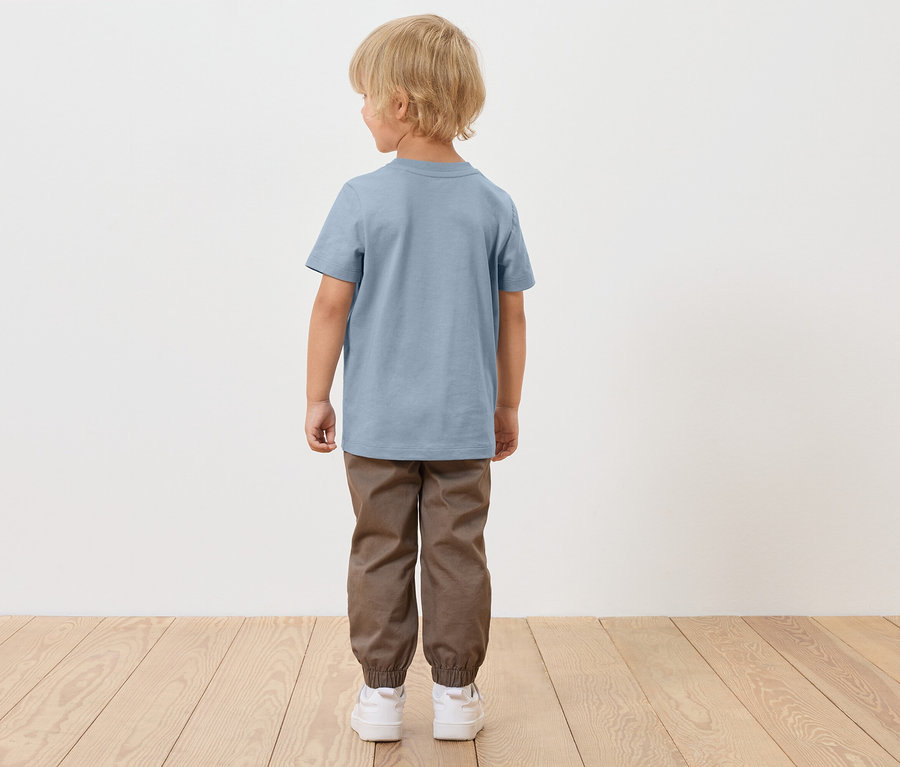 Un garçon blond, vu de dos, porte un t-shirt bleu, un pantalon marron et des chaussures blanches.