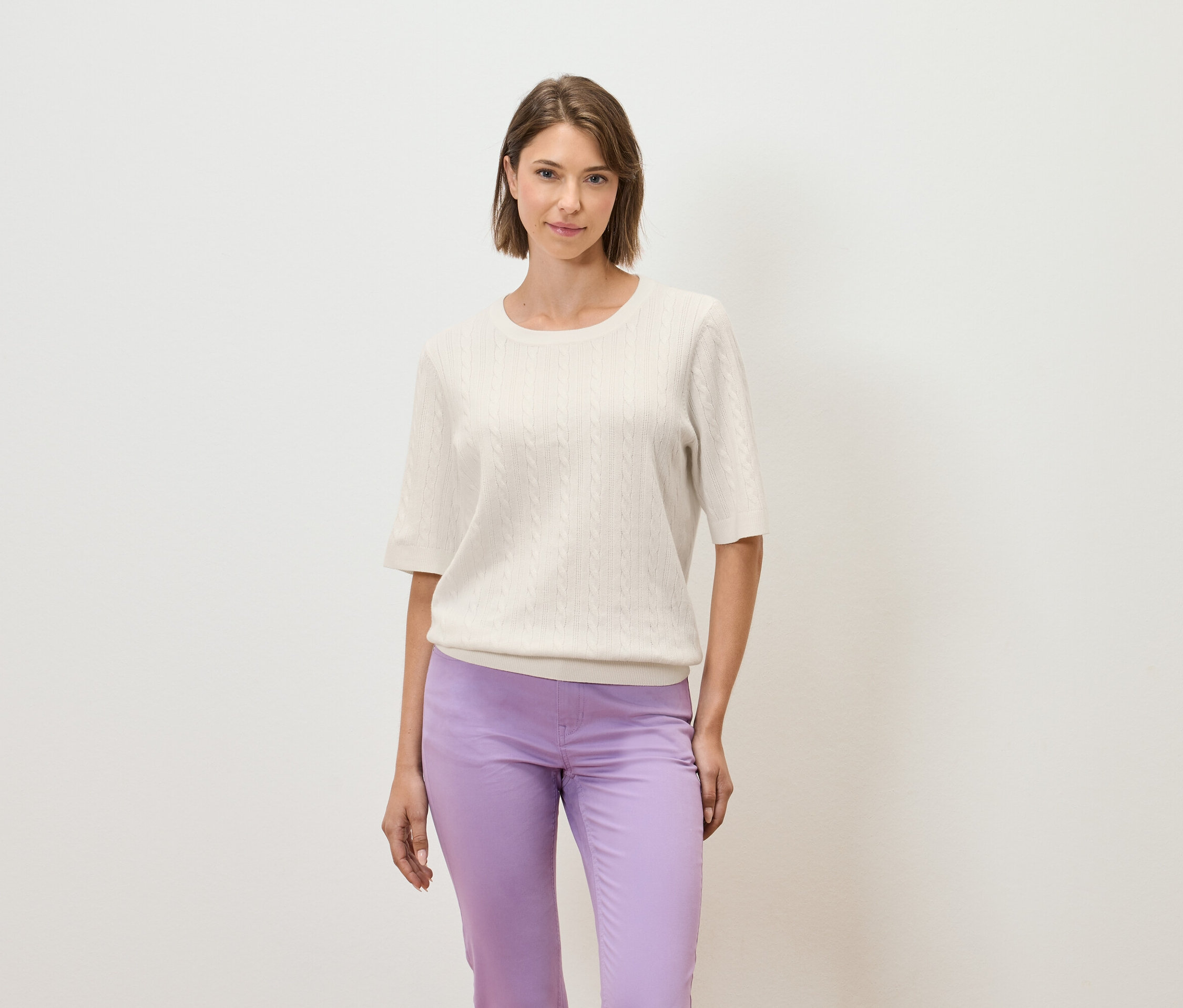 Une femme pose sur un fond clair, portant un pull crème à manches courtes et un pantalon violet.