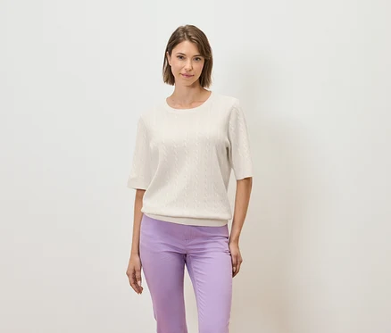 Une femme pose sur un fond clair, portant un pull crème à manches courtes et un pantalon violet.