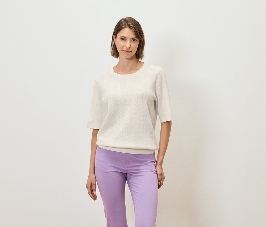 Une femme pose sur un fond clair, portant un pull crème à manches courtes et un pantalon violet.