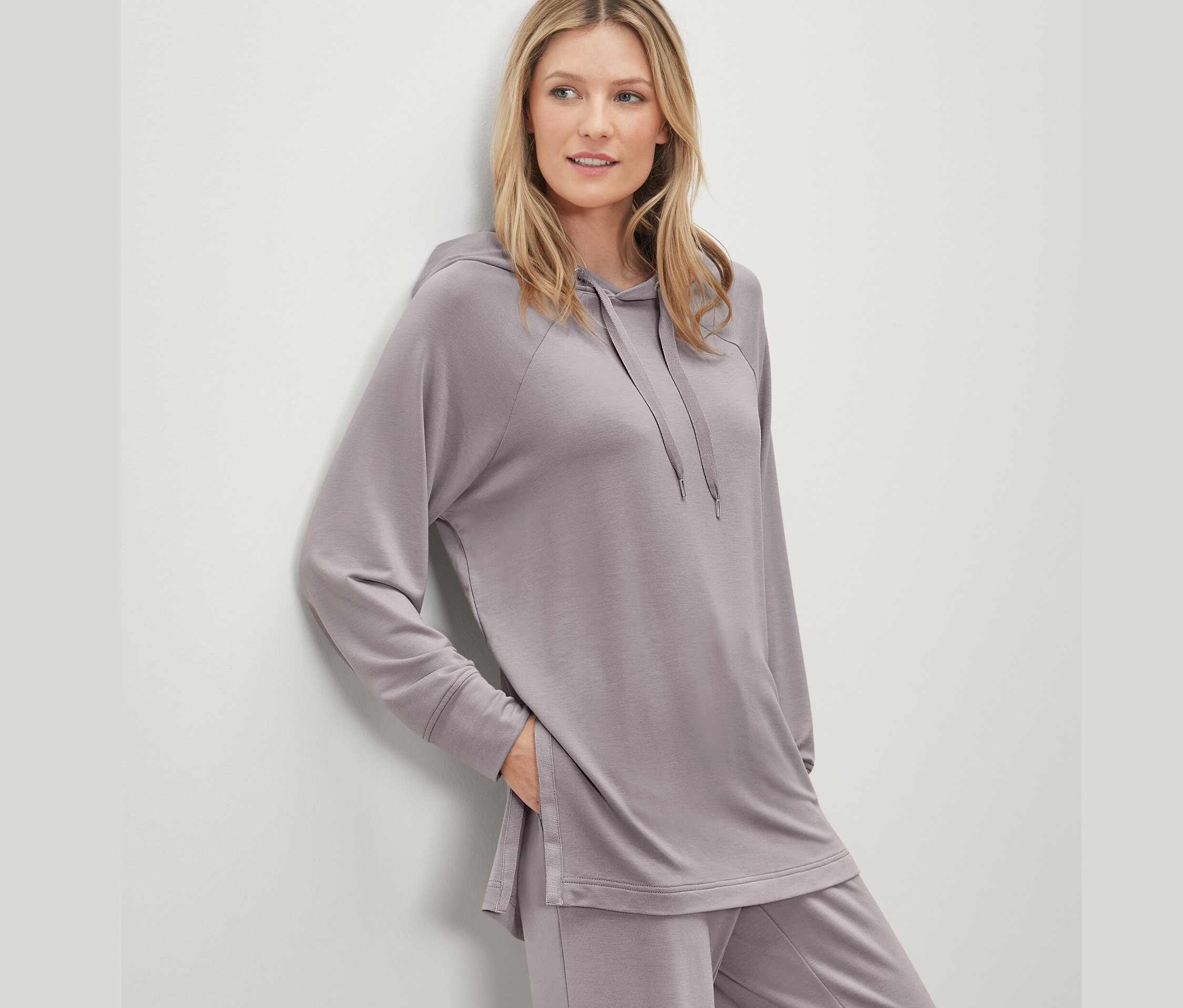 Femme portant un ensemble gris avec un pull à capuche d’intérieur, appuyée contre le mur.