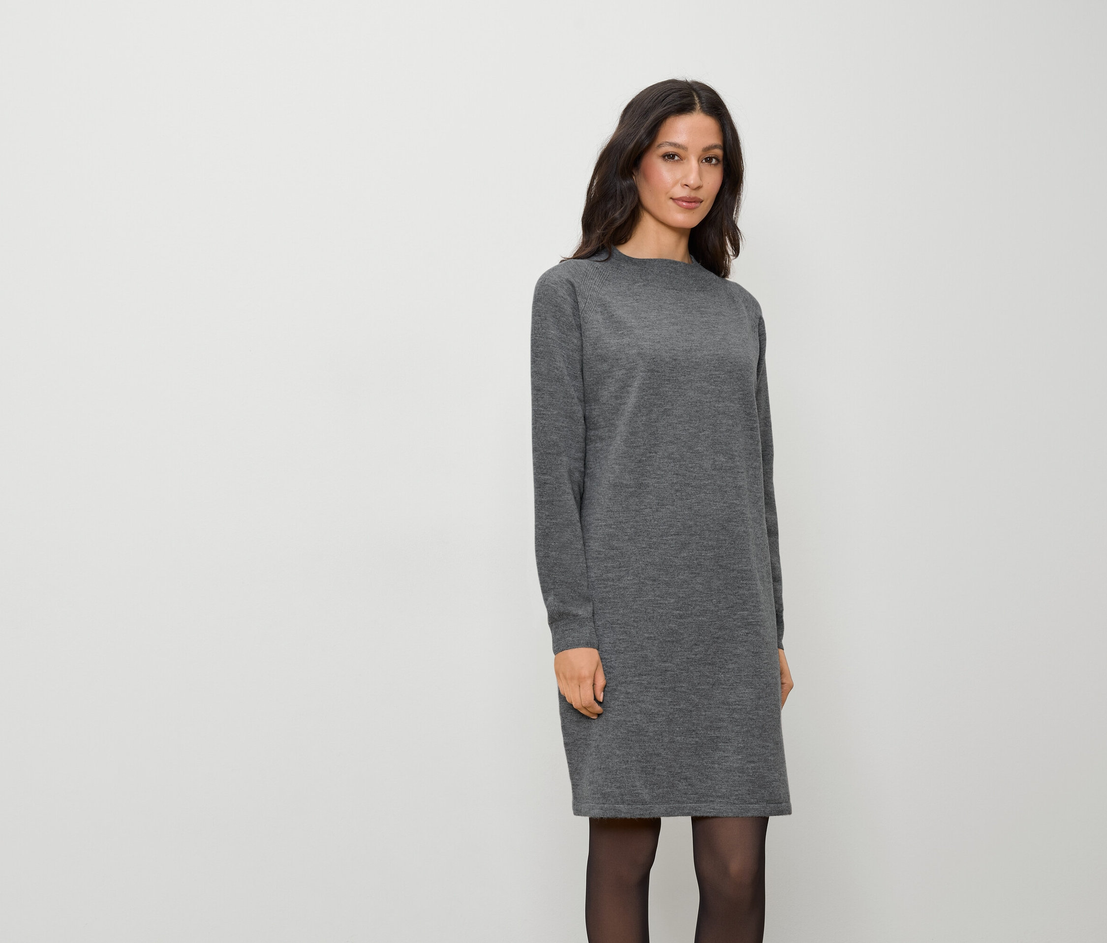 Une femme portant une robe grise à manches longues et des collants noirs se tient sur un fond blanc.