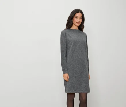 Une femme portant une robe grise à manches longues et des collants noirs se tient sur un fond blanc.