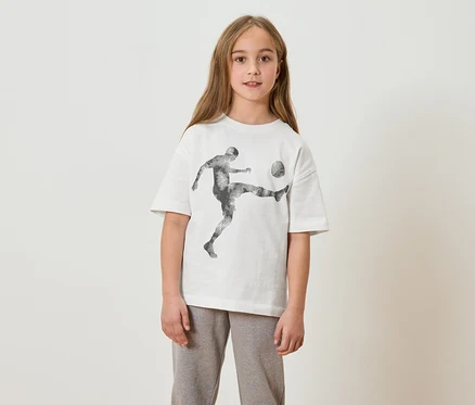 Une fille aux cheveux longs porte un t-shirt blanc avec un imprimé de joueur de foot et un pantalon de jogging gris.