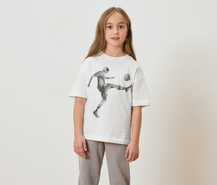 Une fille aux cheveux longs porte un t-shirt blanc avec un imprimé de joueur de foot et un pantalon de jogging gris.