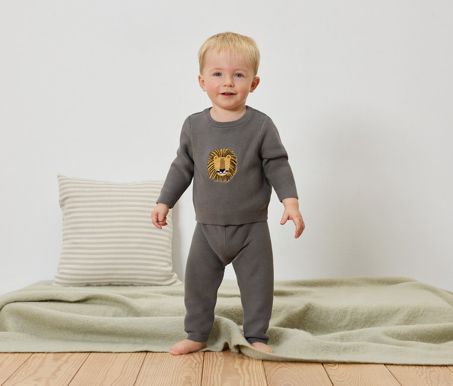 Un jeune garçon se tient pieds nus sur une couverture vert clair, vêtu d'un pull gris avec un motif de lion et d'un pantalon gris.