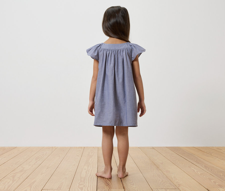 Vue de dos d'une jeune fille aux cheveux foncés portant une robe bleue et debout pieds nus sur un plancher en bois devant un mur blanc.