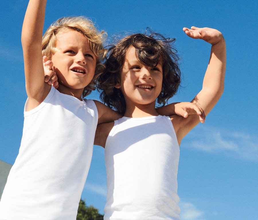Deux enfants souriants aux cheveux bouclés blonds et bruns s'embrassent devant un ciel bleu éclatant, vêtus de débardeurs blancs.