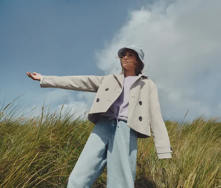 Une jeune femme se tient dans un champ, portant un trench-coat marron clair, un sweat-shirt violet, un jean et un chapeau de pêcheur bleu.