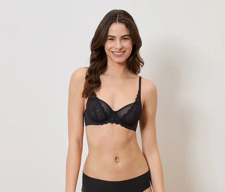 Une femme souriante aux cheveux bruns ondulés porte un soutien-gorge en dentelle noire et une culotte noire sur fond blanc.