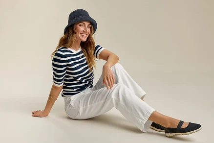 Femme assise portant un jeans grand confort, un t-shirt rayé et un chapeau. Elle sourit.