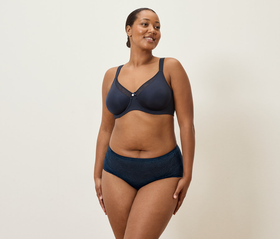 Une femme d'âge moyen aux cheveux foncés et aux sous-vêtements bleu marine se tient devant un fond crème.