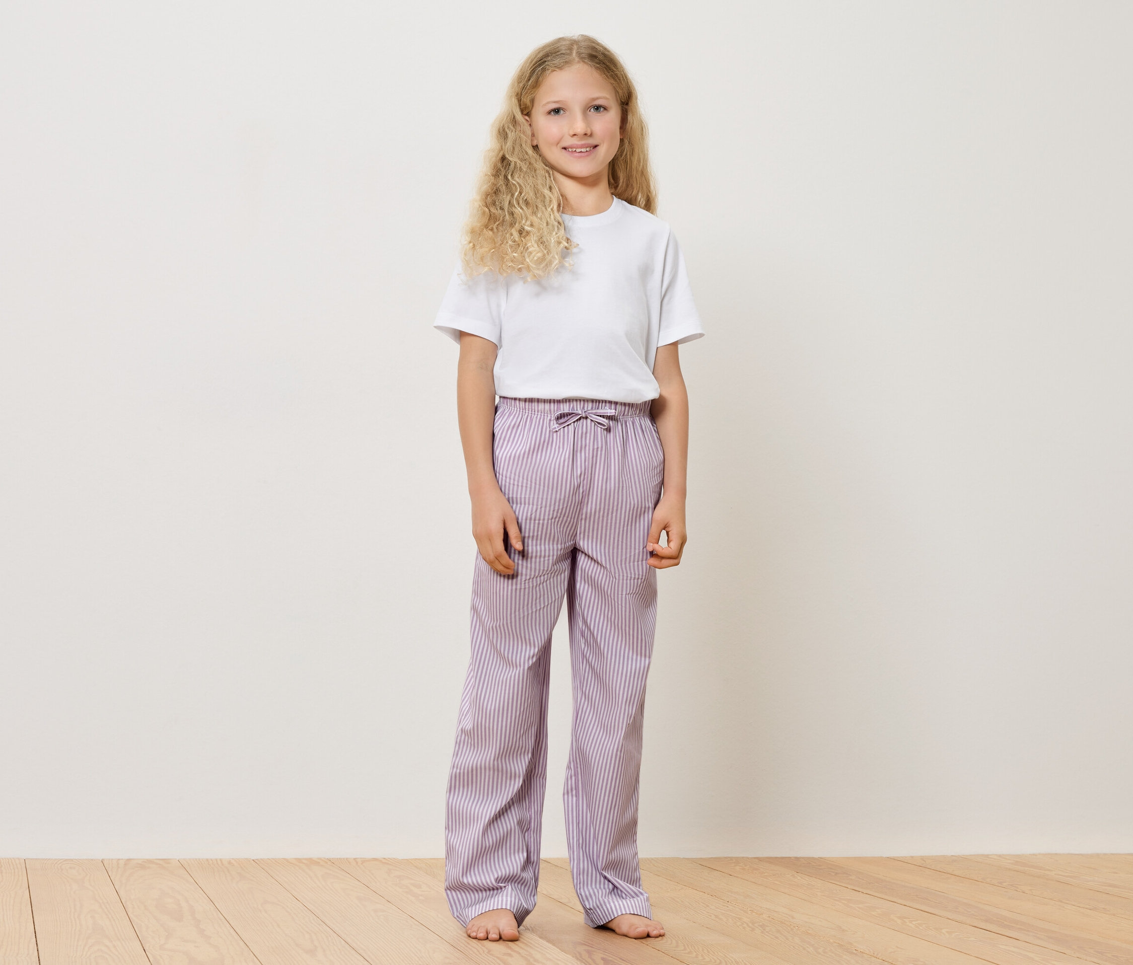 Une jeune fille aux cheveux blonds et bouclés, portant un t-shirt blanc et un pantalon rayé violet, se tient pieds nus sur un plancher en bois devant un mur blanc.