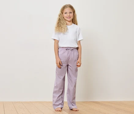 Une jeune fille aux cheveux blonds et bouclés, portant un t-shirt blanc et un pantalon rayé violet, se tient pieds nus sur un plancher en bois devant un mur blanc.