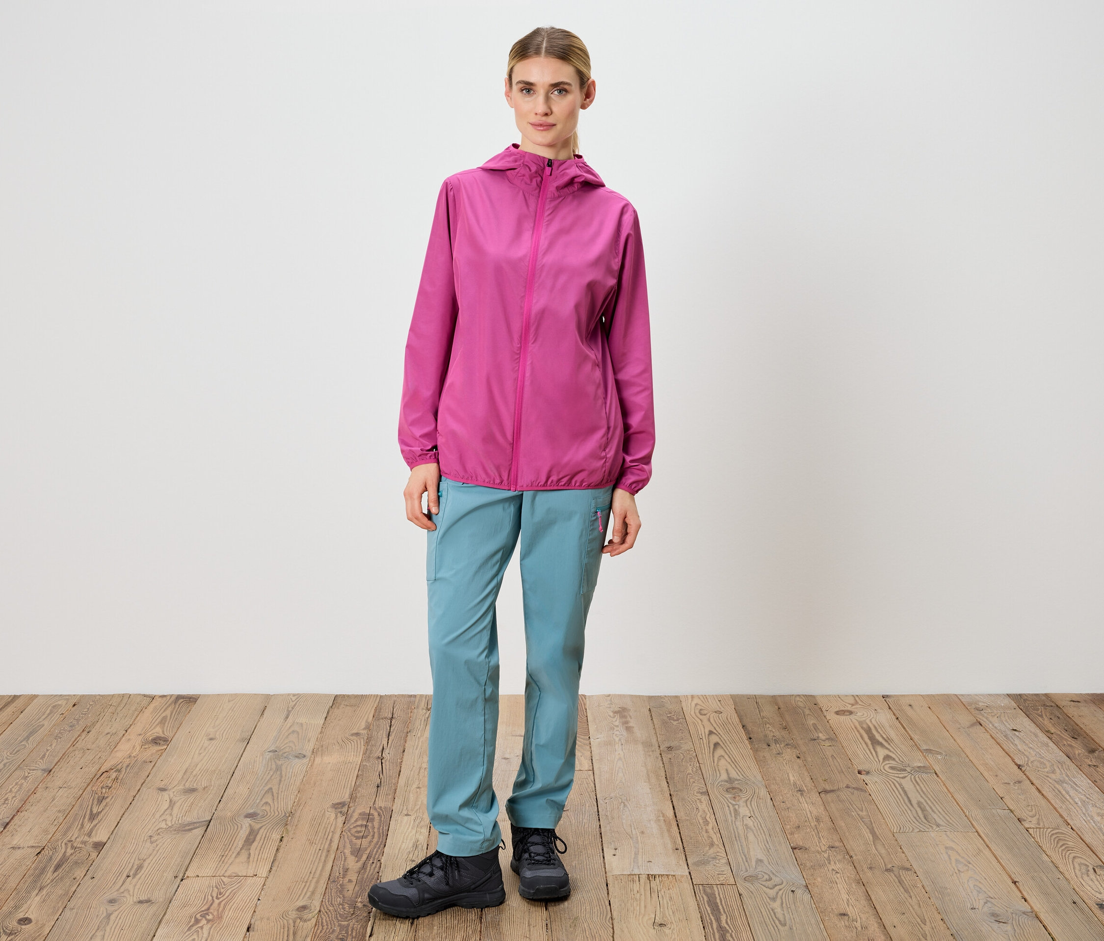 Une femme se tient devant un mur blanc, portant une veste rose, un pantalon bleu et des chaussures noires.