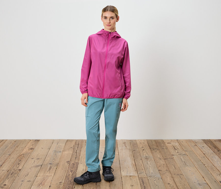 Une femme se tient devant un mur blanc, portant une veste rose, un pantalon bleu et des chaussures noires.