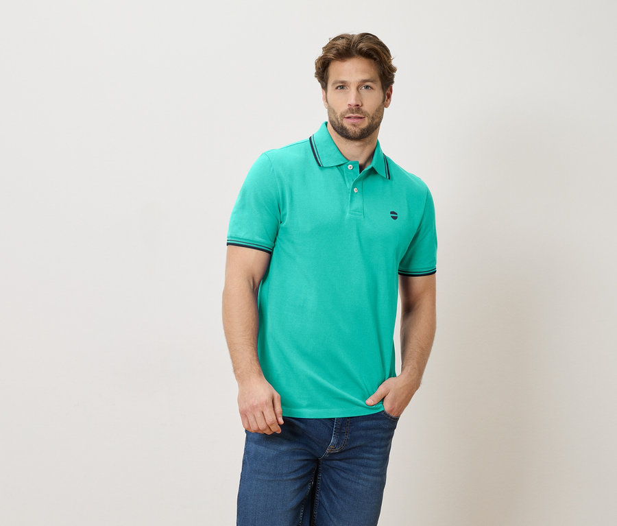 Homme portant un polo vert émeraude et un jean, une main dans la poche.