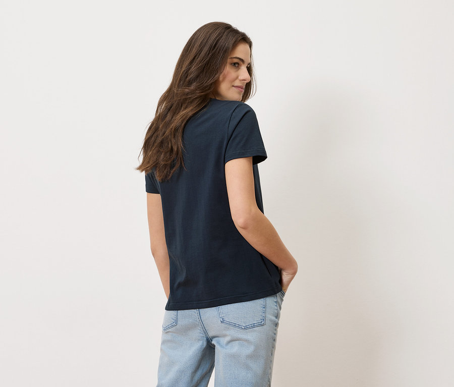 Une femme portant un t-shirt foncé et un jean bleu clair se tient devant un mur blanc.