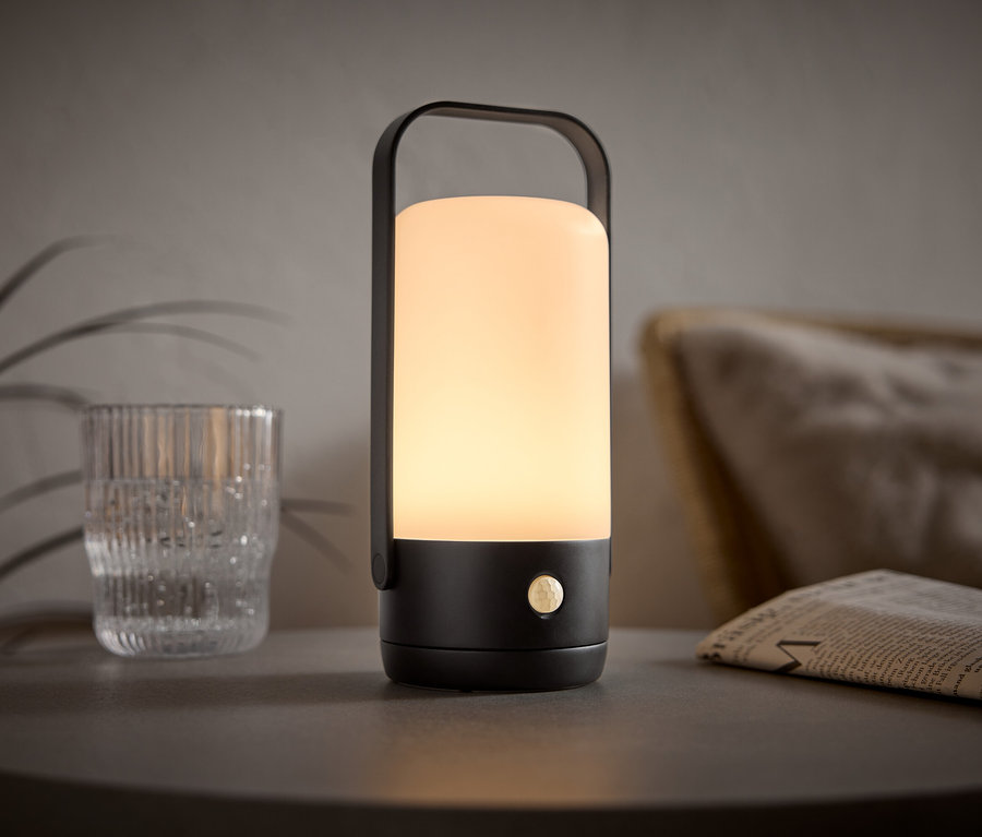 Une lampe portable éclairée avec une poignée noire est posée sur une table à côté d'un verre et d'un journal.