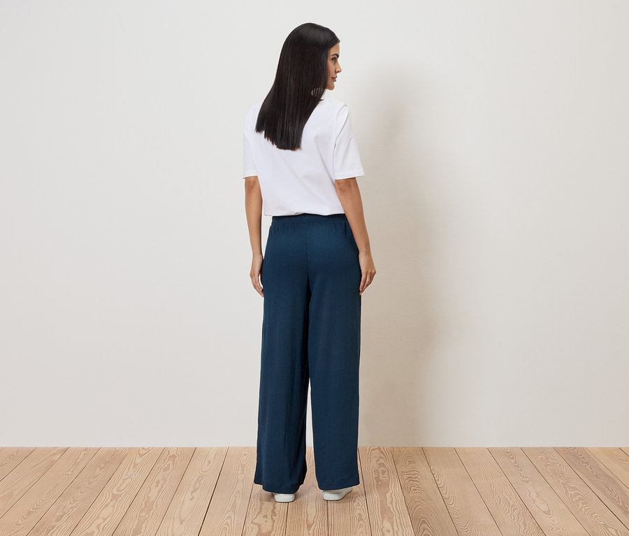 Une femme aux cheveux foncés, vue de dos, porte un t-shirt blanc et un pantalon ample bleu foncé devant un mur blanc.