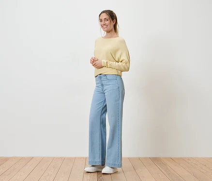 Une femme souriante, les cheveux attachés, porte un pull jaune, un jean bleu clair et des chaussures blanches. Elle se tient sur un plancher en bois devant un mur blanc.