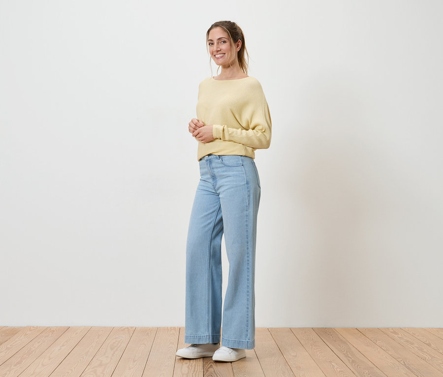 Une femme souriante, les cheveux attachés, porte un pull jaune, un jean bleu clair et des chaussures blanches. Elle se tient sur un plancher en bois devant un mur blanc.