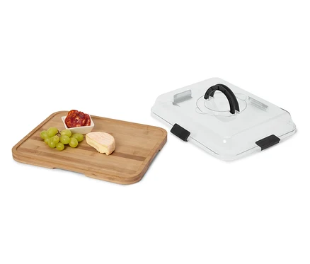Fromage, saucisson et raisins sur une planche en bois à côté d'un couvercle de service transparent avec une poignée noire.