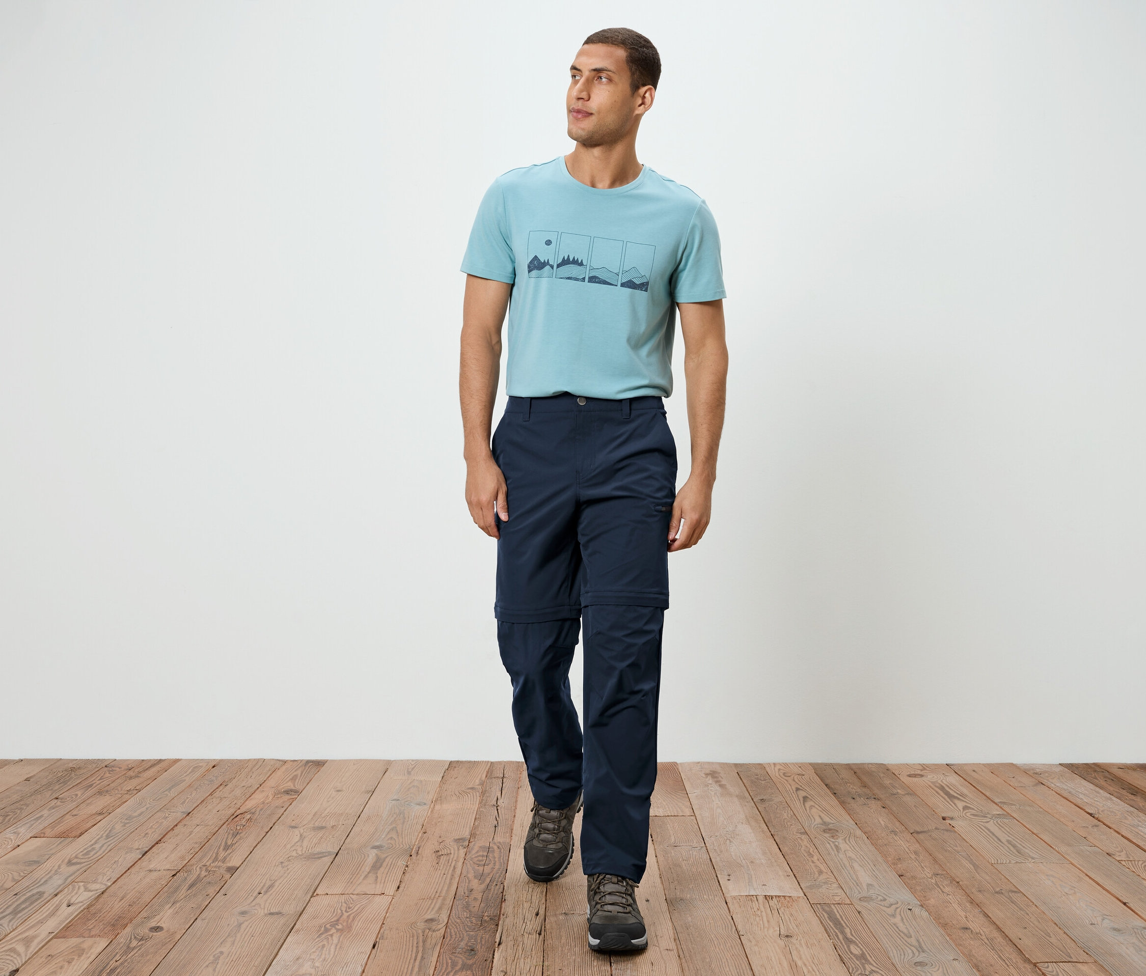 Un homme marche sur un plancher en bois, il porte un t-shirt bleu, un pantalon bleu marine et des baskets grises.